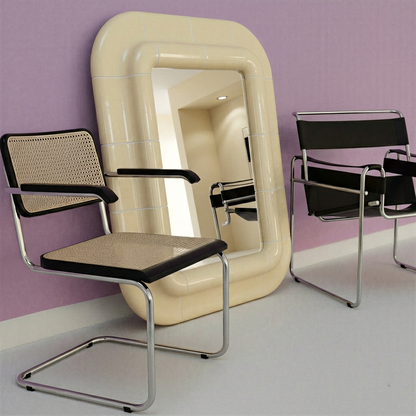 Soft butter glossy mirror with rounded tile frame, leaning against a lavender wall between modern tubular chairs.