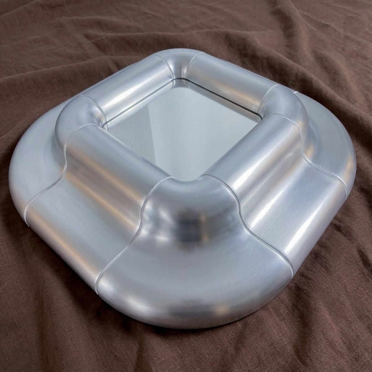 Lolo mirror in Steel finish lying on brown fabric, its rounded metallic volumes catching soft reflections